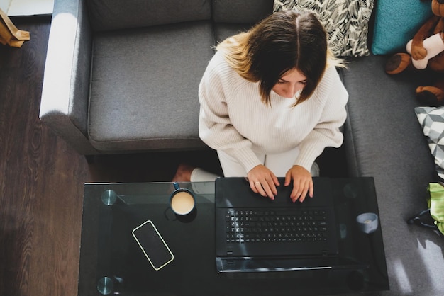 Schöne und ernsthafte junge Frau, die mit Stofftieren auf dem Sofa sitzt und zu Hause mit einem Laptop bei einer Tasse Kaffee arbeitet, das Konzept der Fernarbeit zu Hause