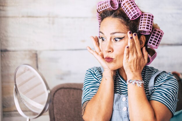 Schöne und lustige kaukasische erwachsene Frau, die sich zu Hause vor dem Spiegel mit Make-up im Gesicht fertig macht - Falten überprüfen und älteres Konzept für junge Leute bekommen - Lockenwickler auf Haaren