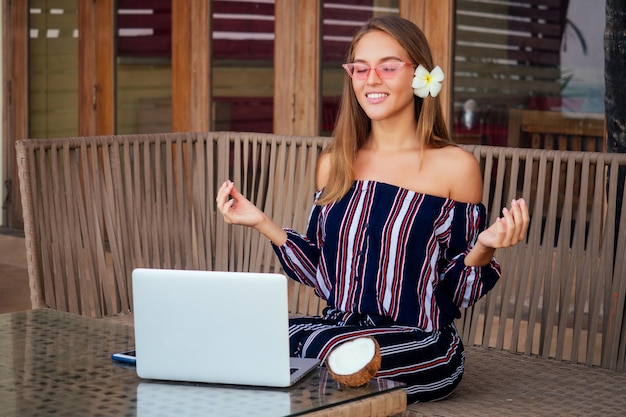 Schöne und ruhige Geschäftsfrau, die an einem Tisch mit einem Laptop in einem Sommercafé in der Lotusposition sitzt und meditiert und sich entspannt