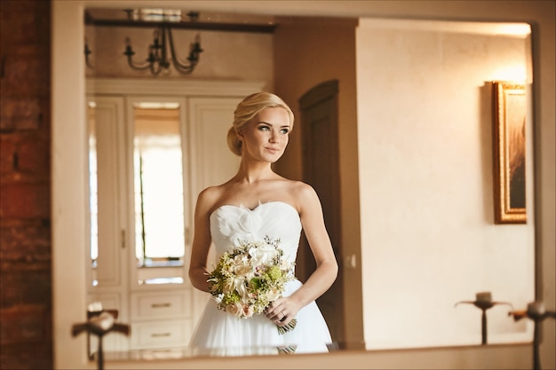 Schöne und sinnliche junge Braut, sexy blondes Modellmädchen mit charmantem Lächeln und mit Blumenstrauß in ihren Händen im weißen Kleid, das im Innenraum aufwirft, Hochzeitsvorbereitung am Morgen