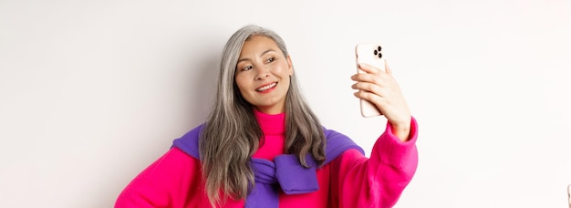 Schöne und stilvolle asiatische Seniorin, die Selfie auf dem Smartphone macht, lächelt und posiert im trendigen Pi