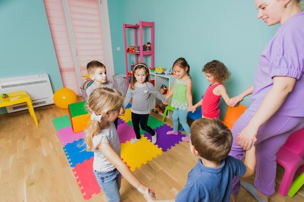 Schöne Vorschullehrerin, die während des Tanzunterrichts mit Kindern im Kreis geht Neue Teambuilding-Techniken lernen Glückliche Kinder mit ihrem jungen Lehrer in der Kindertagesstätte