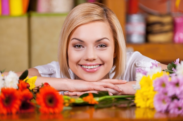 Schöne weibliche Floristin. Schöne junge Frau mit blonden Haaren, die sich voller Blumen am Tisch lehnt und lächelt