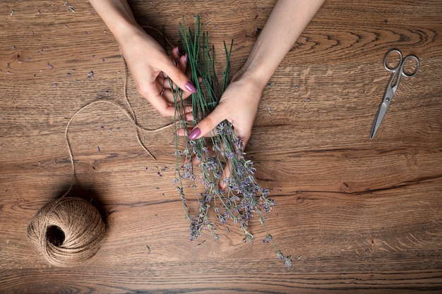 Schöne weibliche Hände binden einen Haufen trockener Lavendel auf dem Hintergrund der Eichentisch-Draufsicht