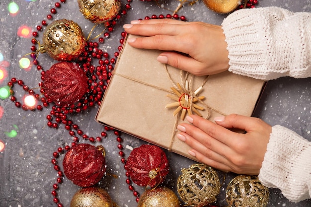 Schöne weibliche Hände der Nahaufnahme mit Maniküre, die Geschenkbox hält Verpackt in Kraftpapier auf grauem Hintergrund mit roten und goldenen Kugeln und Perlen, Draufsicht. Urlaub, Neujahr, Weihnachten, Geburtstag, Winter