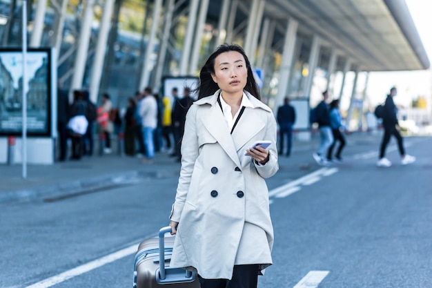 Schöne weibliche Touristin in der Nähe des Flughafens, eine Chinesin, die mit einem großen Koffer spazieren geht, eine asiatische Frau, die ein Telefon hält, die die Anwendung zum Buchen einer Unterkunft und zum Bestellen eines Taxis verwendet