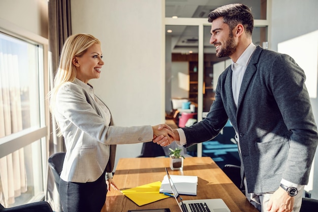 Schöne weibliche und hübsche männliche Geschäftspartner in eleganten Anzügen, die sich die Hände schütteln