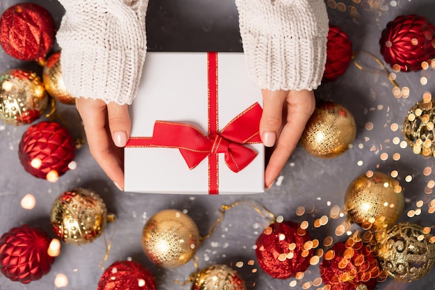 Schöne weiße Geschenkbox in der Hand Mädchen auf grauem Hintergrund mit glänzenden Weihnachtskugeln