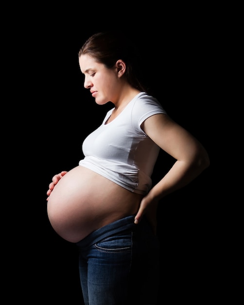 Schöne werdende schwangere Frau, die ihren Bauch über einem schwarzen Hintergrund isoliert sieht looking