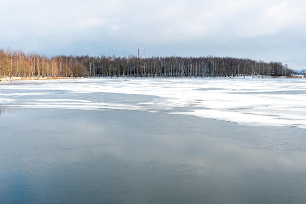 Schöne Winterlandschaft Wald am Seeufer an einem sonnigen frostigen Tag Panorama der Küste mit Schnee und Birkenhain bedeckt Eis auf dem Fluss und die Reflexion der Wolken im Wasser