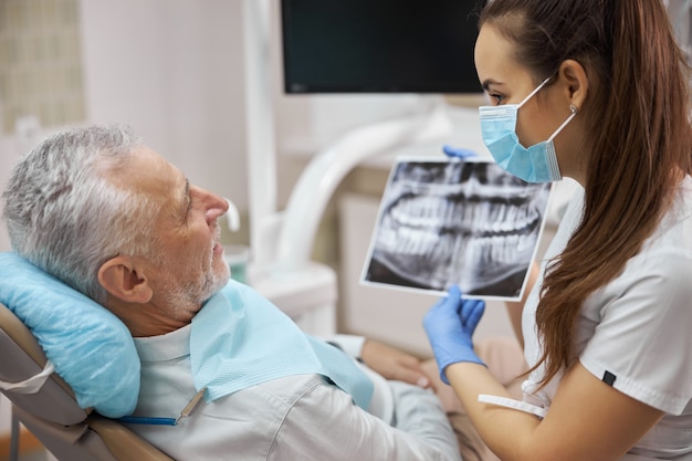 Schöne Zahnärztin, die ältere Patienten anschaut, während sie ihm ein Röntgenbild seiner Zähne zeigt