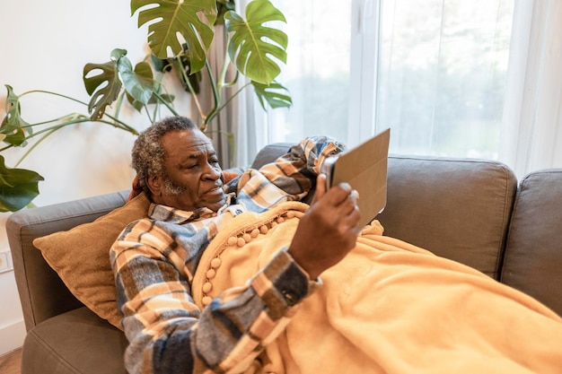 Schöner älterer Mann, der zu Hause auf einem Sofa liegt und mit einem Handheld-Tablet Geschenke für Freunde und Familie online kauft oder Online-Videoanrufe tätigt