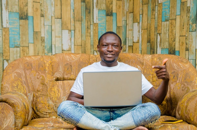 Schöner afrikanischer Mann, der mit Laptop auf dem Sofa im Zimmer sitzt