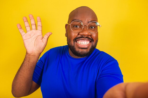 Schöner afro-brasilianischer Mann mit Brille, blauem Hemd über gelbem Hintergrund, der Selfie mit Hallo-Schild-Schild-Gruß-Begrüßungs-Videoanruf-Selfie-interaktives Foto macht