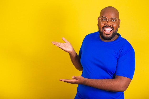 Schöner afro-brasilianischer Mann mit Brille, blauem Hemd über gelbem Hintergrund, mit offenen Händen, der die Seite präsentiert und darauf hinweist, dass der negative Raum für Werbekopien angezeigt wird