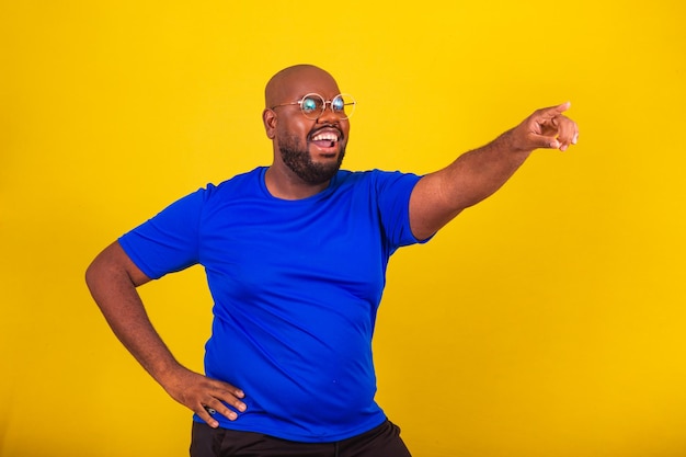 Schöner afro-brasilianischer Mann mit Brille, blauem Hemd über gelbem Hintergrund, Zeigefinger, der dorthin zeigt, was darauf hindeutet, dass er ein Werbeangebot zeigt