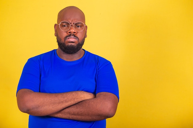 Schöner afro-brasilianischer Mann mit Brille, blaues Hemd über gelbem Hintergrund, Arme verschränkt, mürrisch, nervös, traurig, unglücklich, frustriert