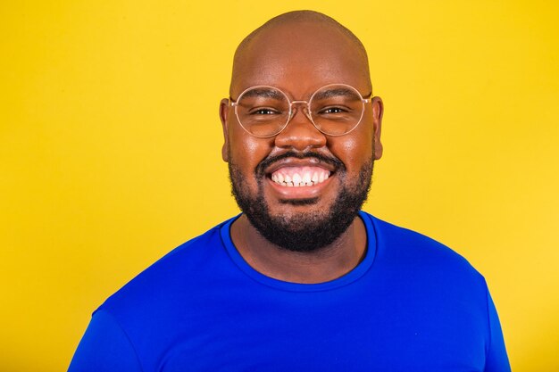 Schöner afro-brasilianischer Mann mit Brille, blaues Hemd über gelbem Hintergrund, Nahaufnahme, Foto, das ein Lächeln-Konzept von Glück, Freude, Frieden, Euphorie zeigt