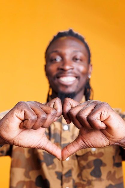 Schöner afroamerikanischer Mann, der im Studio vor gelbem Hintergrund eine romantische Geste macht und mit den Händen eine Herzform macht. Fröhliche, positive Person, die Zuneigung und Emotionen zeigt und Liebeszeichen macht