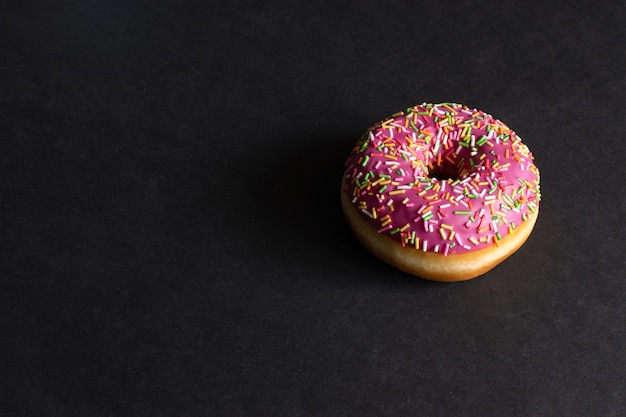 schöner appetitlicher Donut mit rosa Zuckerguss und Gebäckstreuseln auf einem stilvollen schwarzen Hintergrund Foto für Konditoreienmenüs und Werbung