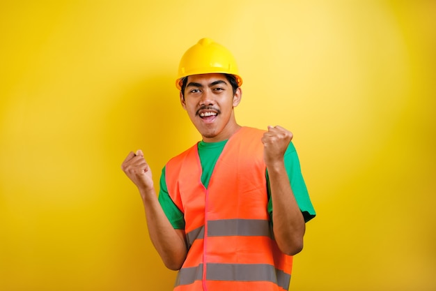 Schöner asiatischer Arbeiter mit Uniform und Helm über isoliertem gelbem Hintergrund, der überrascht und erstaunt über den Erfolg mit erhobenen Armen und offenen Augen feiert. Gewinner-Konzept.