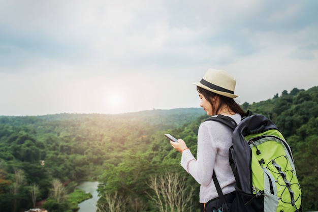 Schöner asiatischer Frauenreisender mit dem Rucksack, der in den Ferien auf Berg mit der Anwendung des Mobiles sich entspannt