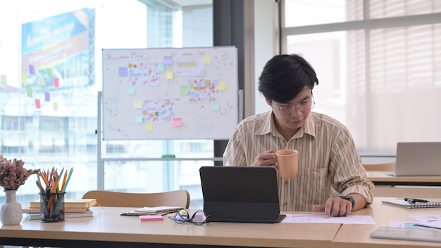 Schöner asiatischer männlicher Manager, der eine Kaffeetasse hält und im modernen Büro arbeitet