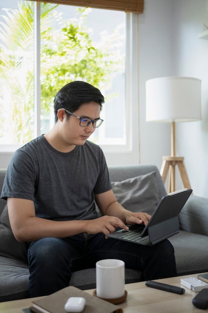 Schöner asiatischer Mann, der auf dem Sofa sitzt und online mit Computertablette arbeitet.