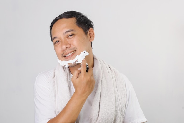 Schöner asiatischer Mann, der sich das Gesicht rasiert und die Kamera anschaut, während er auf grauem Hintergrund steht