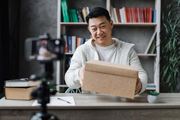 Schöner asiatischer Mann in Freizeitkleidung, der beim Auspacken der Geschenkbox ein Video vor der Kamera aufnimmt