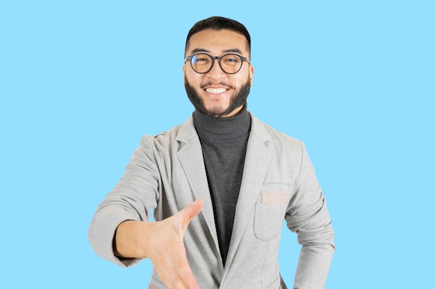 Schöner asiatischer Mann mit Brille und Bart, der eine Businessman39s-Jacke trägt, die Hände mit einer selbstbewussten Geste auf blauem Hintergrund schüttelt