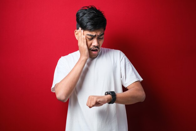 Schöner asiatischer Mann mit weißem T-Shirt, der besorgt auf die Uhr schaut und Angst hat, auf rotem Hintergrund zu spät zu kommen