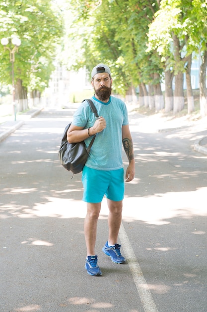 Schöner Athlet. Sporttrainer mit Rucksack bereit für das Training im Freien. Sport ist Lebenseinstellung. Kerl hübsches bärtiges Gesicht in Sportuniform im Park. Sommertraining. Täglicher Spaziergang zum Fitnessstudio. Gesundheitsvorsorge.