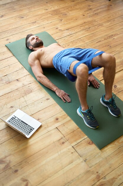 Schöner bärtiger Herr in Shorts, der mit einem Widerstandsband trainiert, während er auf einer Fitnessmatte in der Nähe des Laptops liegt