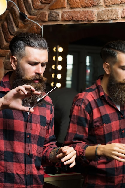 Schöner bärtiger Hipster mit stylischem Haarschnitt und Bart, der eine Schere im rot karierten Hemd in der Nähe des Spiegels mit ernstem Gesicht im Schönheitssalon oder Friseursalon als Friseur in der Nähe der Ziegelwand hält