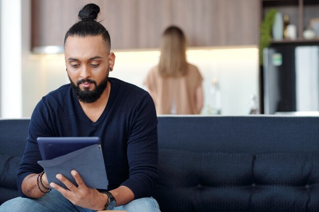 Schöner bärtiger junger Mann, der zu Hause auf dem Sofa sitzt und Artikel auf dem Tablet-Computer liest