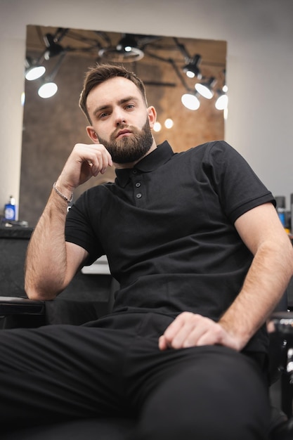Schöner bärtiger männlicher Friseur, der auf einem Stuhl im Friseursalon sitzt
