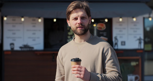 Schöner bärtiger Mann, der eine Tasse Kaffee zum Mitnehmen hält und an einem schönen Tag auf der Straße lächelt und Kaffee trinkt