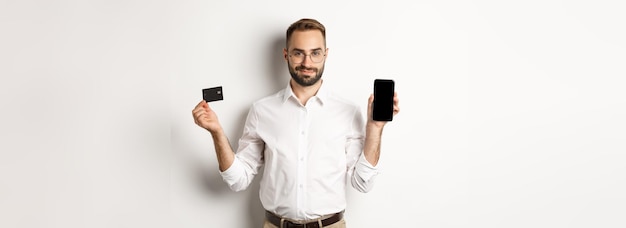 Schöner bärtiger Mann, der Handy- und Kreditkarten-Shopping online zeigt, stehend über weißem Backgr