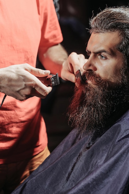 Schöner bärtiger Mann Hipster Brünette mit Bart und Schnurrbart hat Haarschnitt oder Haarschneidemaschine im Friseursalon oder Friseursalon Friseur arbeitet mit Haarschneidemaschine und Kamm