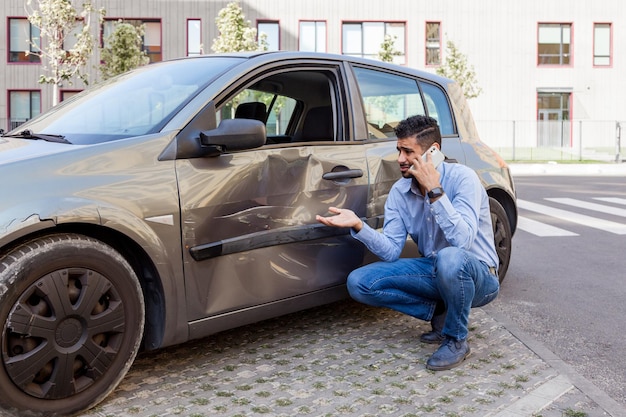 Schöner bärtiger Mann in Jeans und blauem Hemd mit traurigem Gesichtsausdruck, Telefon, Anruf bei der Polizei oder Versicherungsinspektion, beschädigte Autotür mit Dellen und Kratzern. Außenaufnahme.