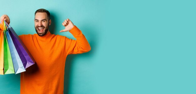 Schöner bärtiger Mann in orangefarbenem Pullover, der Geschenke kauft, auf sich selbst zeigt und Einkaufstüten st zeigt