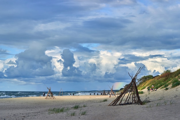 Schöner bewölkter Himmel mit Regenbogen über Sandstrand mit Wigwams. Menschen, die sich am Meer ausruhen