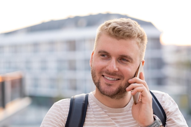 Schöner blonder bärtiger lächelnder Mann mit lässiger Kleidung, der mit dem Smartphone in der Stadt im Freien anruft. Glücklicher Kaukasier mit Rucksack, der per Telefon spricht