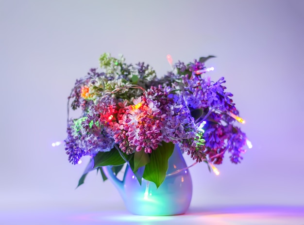 Schöner Blumenstrauß aus duftenden lila Blumen in blauer Keramikvase auf hellem Hintergrund mit bunten Lichtern der festlichen Girlande. Syringa vulgaris oder Fliederpflanze.