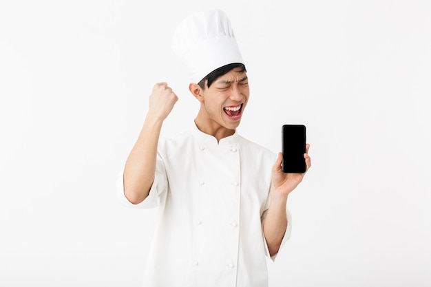 schöner chinesischer Hauptmann in weißer Kochuniform und Kochmütze, die Handy lokalisiert über weißer Wand hält