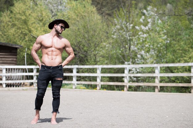 Schöner Cowboy-Mann, der stark auf der Ranch im Freien posiert und schwarze Jeans und Cowboyhut trägt