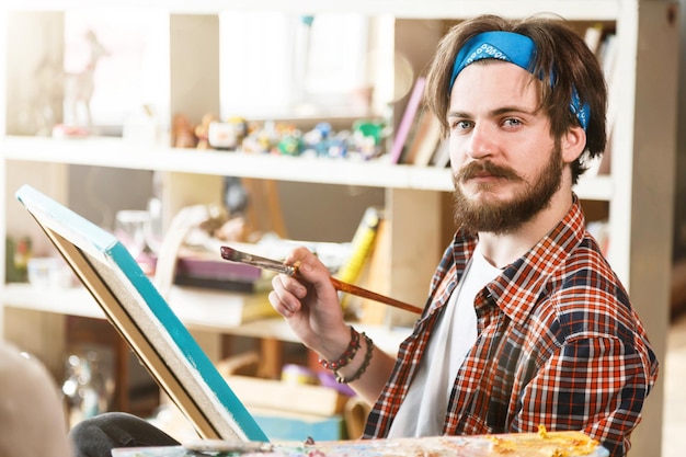 Schöner, dunkelhaariger, bärtiger Hipster-Künstler in blauem Bandana, der in seinem hellen, modernen Studio sitzt und Pinsel hält