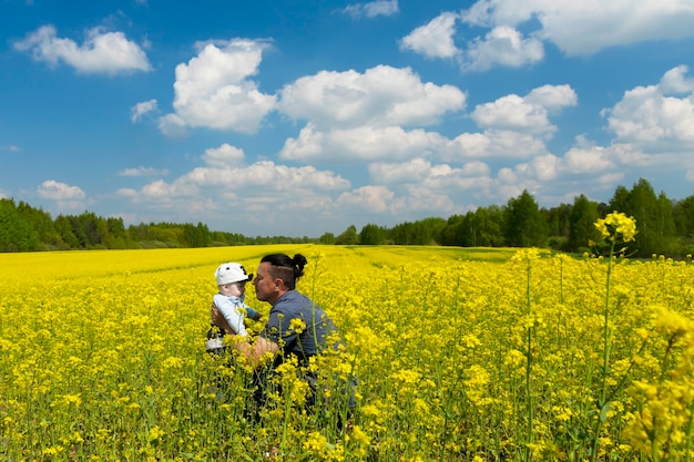 Schöner dunkelhaariger Mann, Papa hält seinen kleinen Sohn in den Armen auf einem blühenden Rapsfeld.