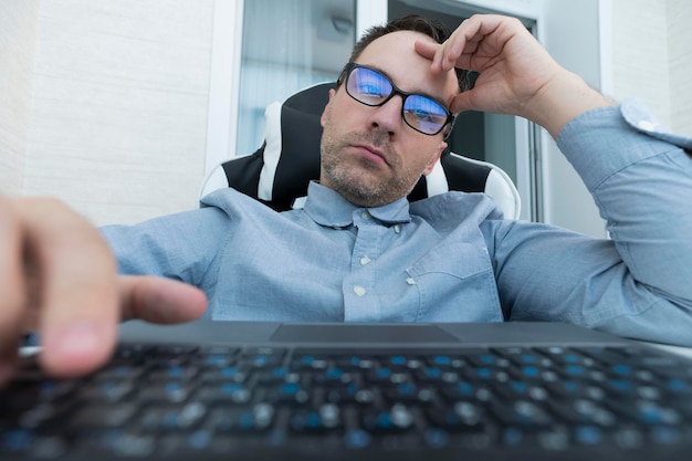 Schöner, ernster Mann, der am Schreibtisch mit Tastatur und Brille an der weißen Wand im modernen Büro sitzt